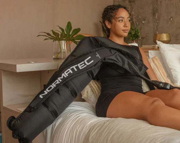 Photo of female wearing a black outfit sitting upright on a bed with the Normatec arm attachments on her right arm inflated.