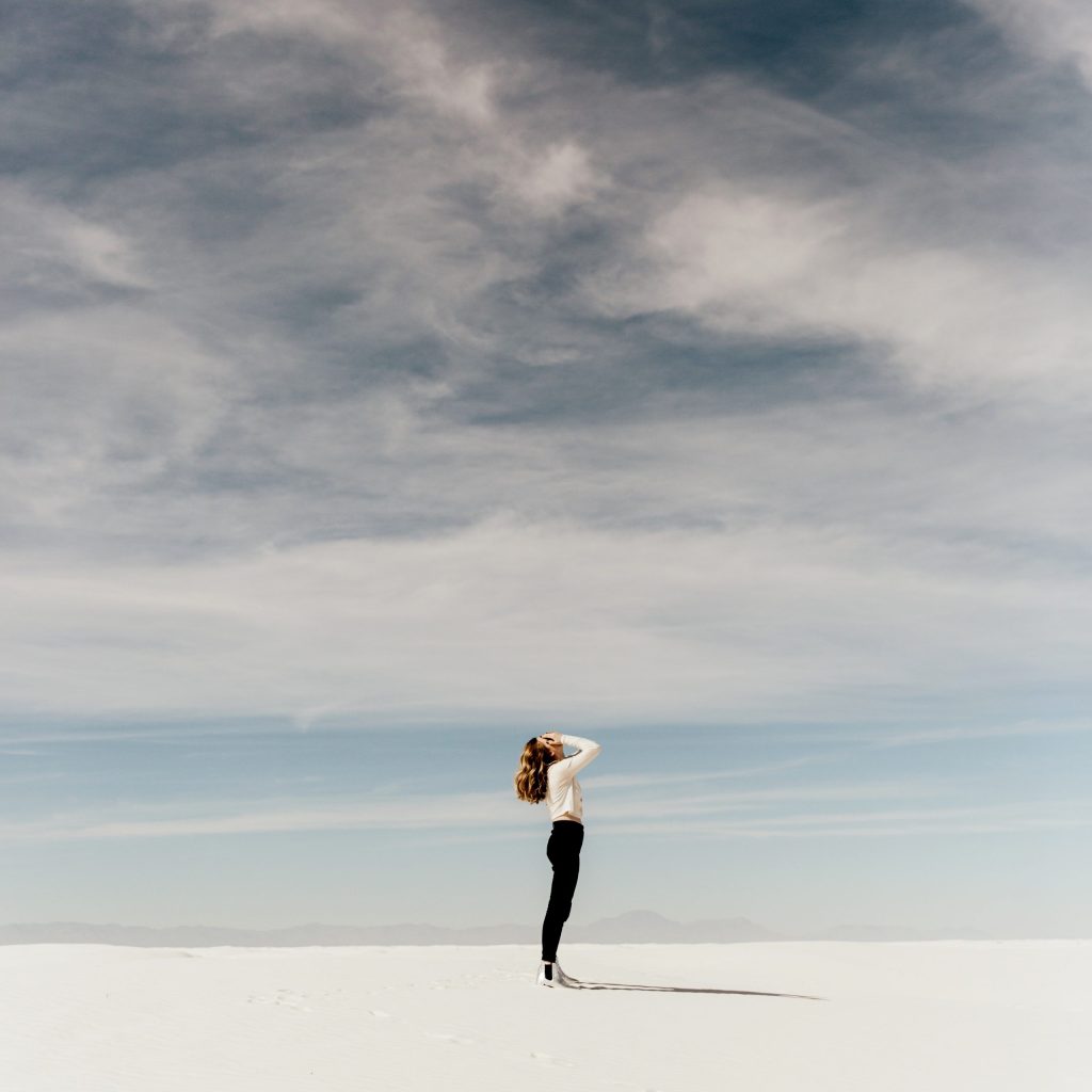 Photo of a female with long hair standing on a beach with her hands over her face.